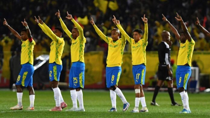 Mamelodi Sundowns players celebrate on the pitch, raising their fingers in unison in front of a packed crowd of yellow-clad supporters.